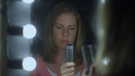 a young woman brushes her hair in mirror and rips out a lock of her hair in shock and sadness