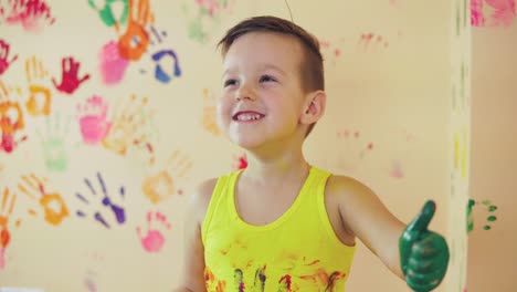 vista de cerca de un niño lindo con manos pintadas después de dejar sus coloridas huellas de manos en la pared y mostrando su pulgar hacia arriba. joven familia feliz. concepto de madre e hijo