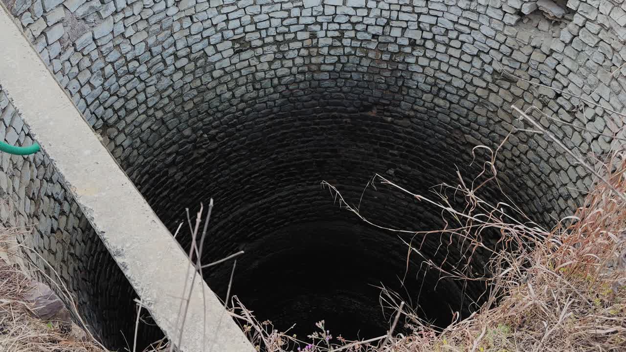 Kamerafahrt auf einen tiefen, mit Stein ausgekleideten Brunnen in einem ländlichen Feld zu, kreisförmige Ziegelwände, die in die Dunkelheit absteigen, trockenes Gras und ein Betonrand deuten auf eine landwirtschaftliche Wasserquelle hin