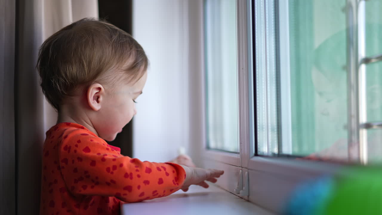 Beautiful little toddler banging cheerfully by the window-sill. Adorable baby looking attentively and curiously at the window.