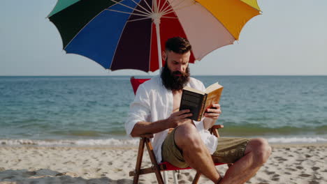 bearded man reading a book on the beach under a colorful umbrella