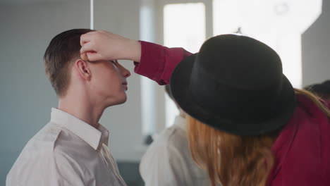 makeup artist wearing black hat and red jacket applies foundation on male profile in bright studio with mirror reflection and professional cosmetic session capturing color blending and skin smoothing