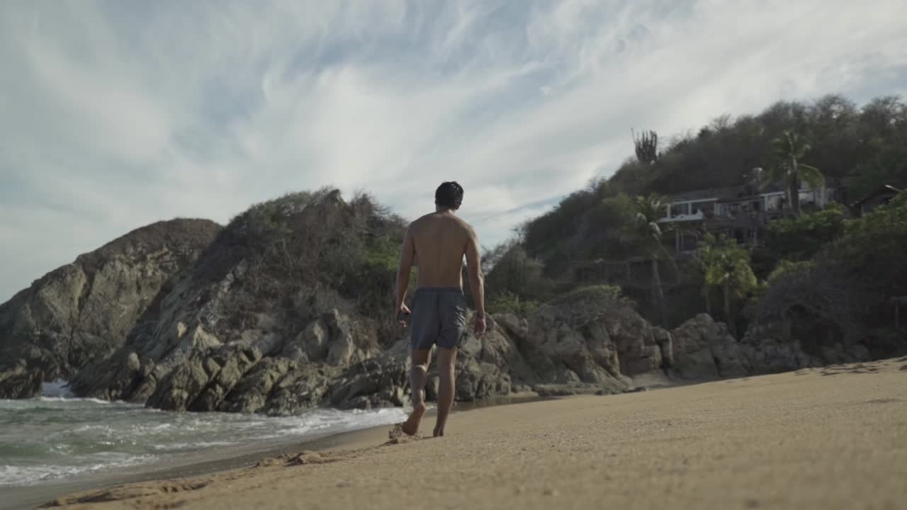A low-angle, wide shot captures a man in swim shorts walking away from the camera and along the water's edge on a sandy beach