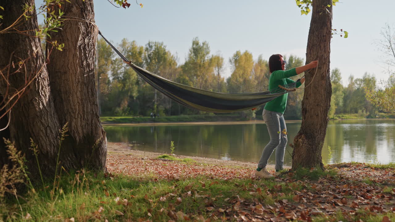 Mujer colgando una hamaca entre árboles junto al lago, asegurando las correas y retrocediendo para probar la tensión, la campista de la tarde ajusta los puntos de amarre bajo la luz solar moteada, orilla frondosa, reflejo tranquilo del bosque.