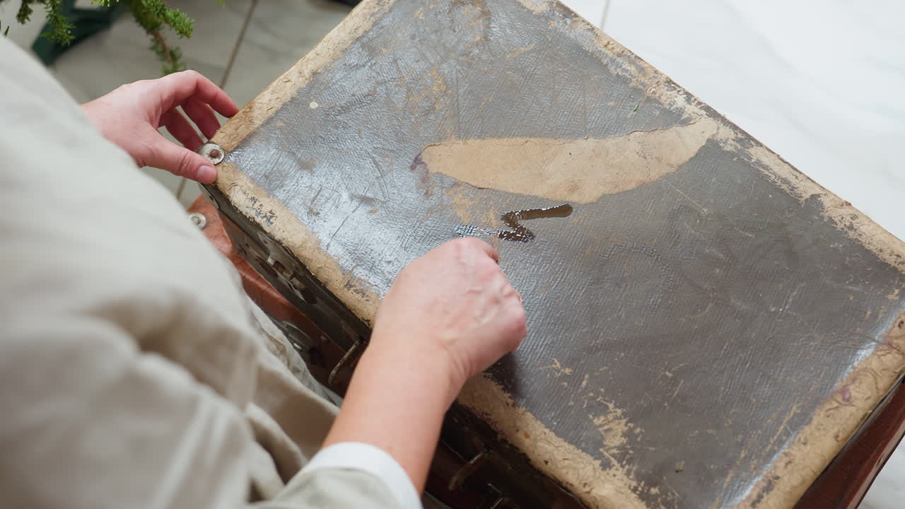 Partial view of woman sketching playful shapes on dusty vintage box surface with finger, creating tactile creative moment during cozy festive indoor setting surrounded by soft seasonal decorations