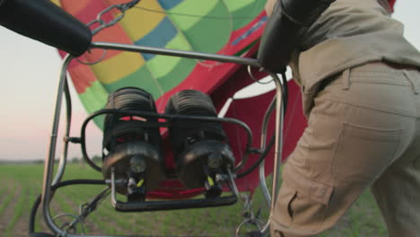 rear view of man operating burner system beside vibrant hot air balloon during setup on grassy field under soft sunset sky with visible tubing, gauges