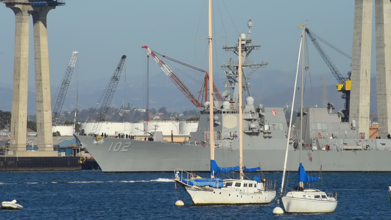 Large naval ship docked near the Coronado Bridge with sailboats and cranes in the harbor