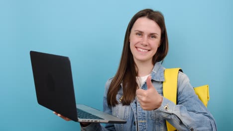 retrato de una hermosa chica adolescente estudiante usar chaqueta de denim y mochila amarilla sostener uso tipo de trabajo en computadora portátil, mostrando el pulgar hacia arriba, fondo azul aislado. educación en el concepto de universidad