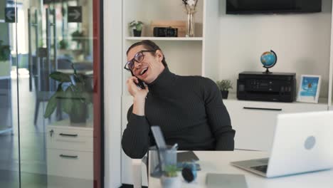 A-man-in-glasses-sits-at-a-table-and-talks-on-the-phone,-occasionally-laughing-and-enjoying-the-conversation