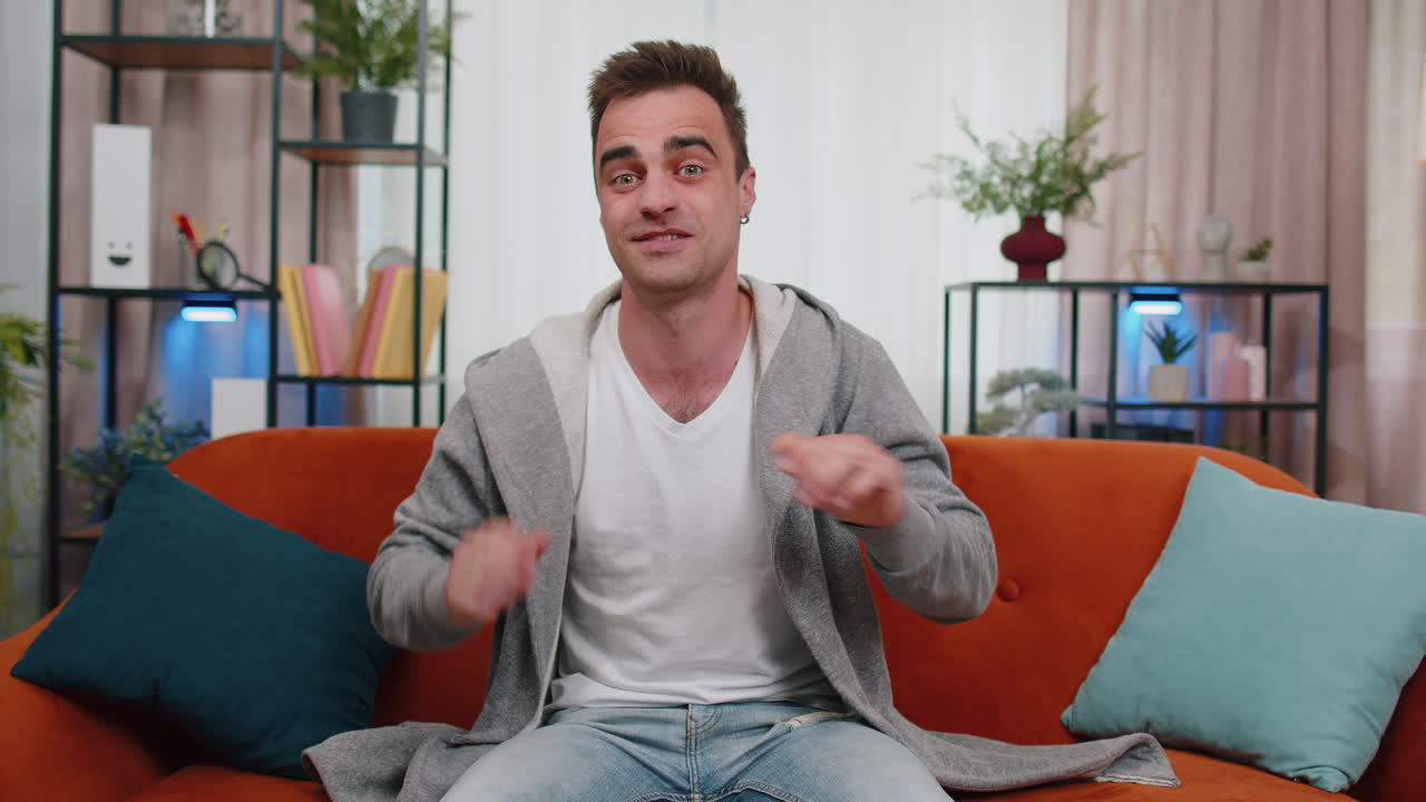 joven sonriendo amistosamente a la cámara, agitando las manos gestando hola, hola, saludando en casa en el sofá