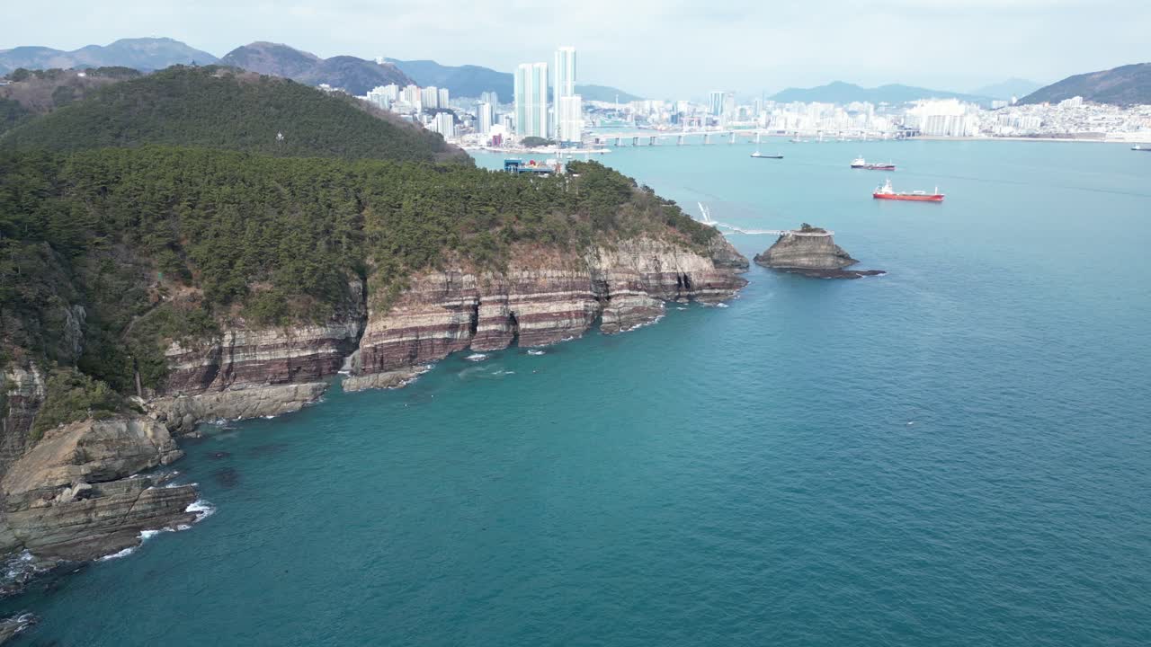 Drone aerial view in South Korea countryside flying over the clear blue sea of Busan next to a green mountain, ships and city in the background sunny day