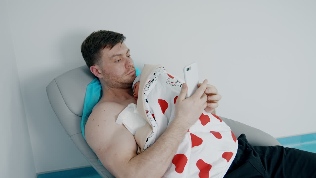 Caucasian man holds his newborn child on the chest. Father is looking at phone while baby is resting.