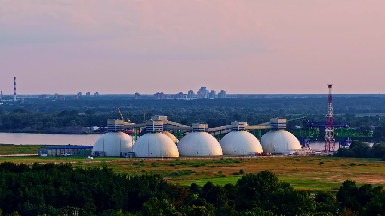 A wide aerial trucking shows the large, dome-shaped silos of the Riga Fertilizer Terminal used for storing dry bulk cargo, situated in the industrial port area of Riga at a stunning sunset, Latvia