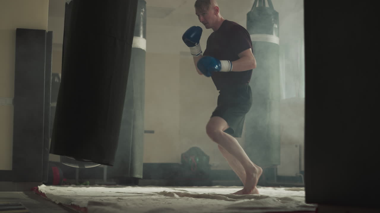 Kick-boxer kicks jumping punching bag in gym. Skilled athlete shows different fighting techniques in gym slow motion. Active combat training in smoke