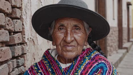 elderly woman with grey hair and wrinkles, wearing a colorful patterned poncho and a wide brimmed hat, smiles gently against a brick wall backdrop