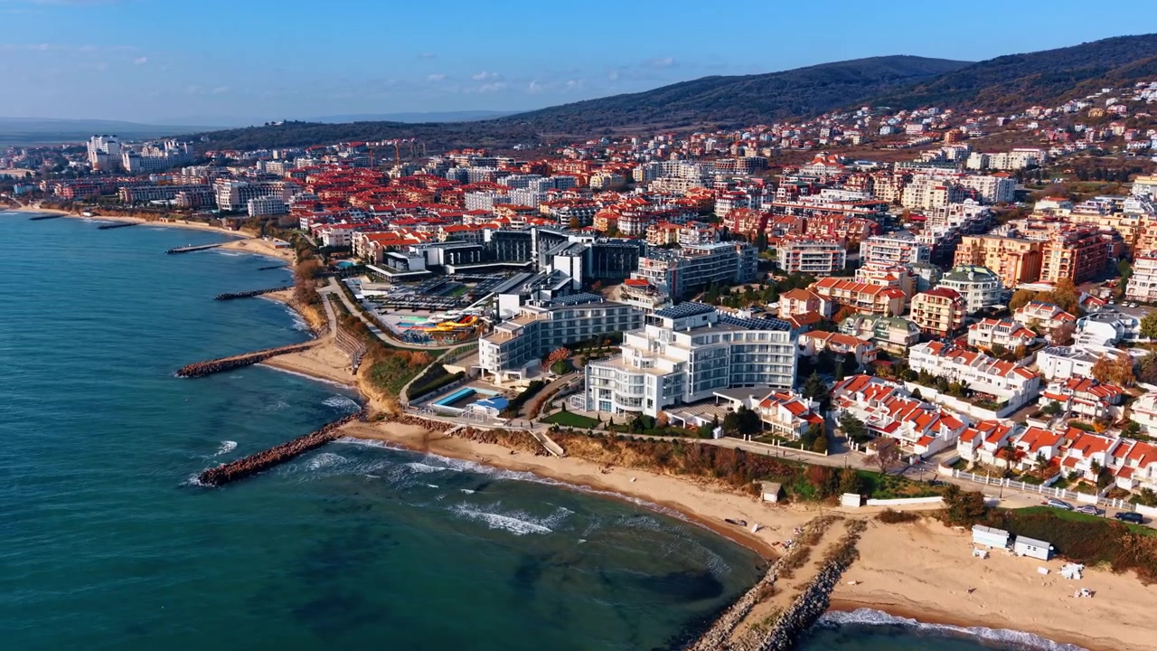 Vista aerea di una città turistica sul lungomare con tetti rossi, piscine e una collina autunnale. Hotel resort con tetti rossi si affacciano su una spiaggia sabbiosa con moli rocciosi, mentre una collina boscosa autunnale fa da sfondo.