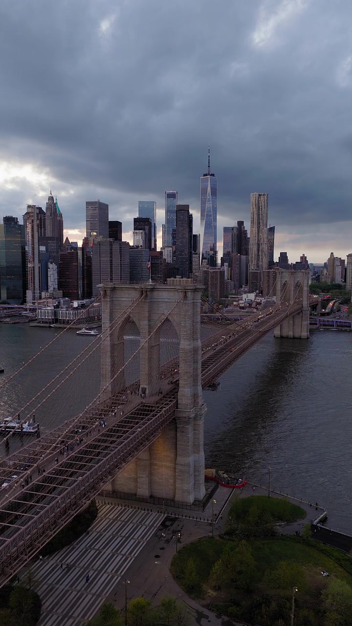 Ponte di Brooklyn, New York City. Ripresa panoramica verticale con drone al tramonto. Bandiera americana sulla torre, cielo nuvoloso dorato. Skyline di Manhattan, con il One World Trade Center visibile.