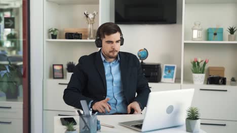 Retrato-De-Un-Hombre-Joven,-Guapo-Y-Alegre,-Escuchando-Música-Con-Auriculares-Inalámbricos-Mientras-Se-Sienta-En-Un-Escritorio-En-Una-Oficina-Luminosa-Con-Atuendo-De-Negocios