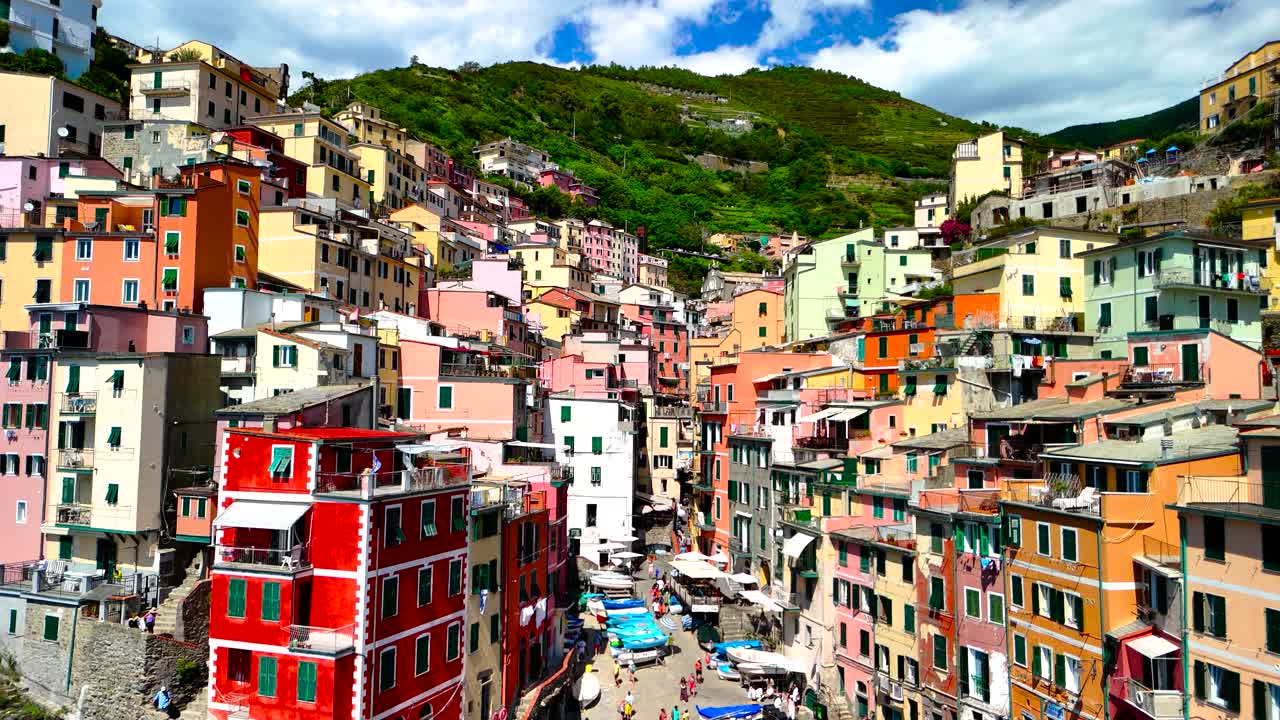 Brightly colored houses stack up the hillside in Riomaggiore. The picturesque village is surrounded by green hills and a beautiful blue sky. Boats rest by the shore below