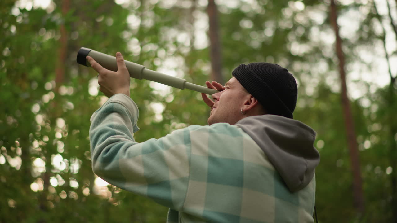Caucasian birdwatcher in dense pine canopy searching for songbirds through homemade spyglass, wearing knit cap and plaid jacket, focused listening and still observation in soft morning light