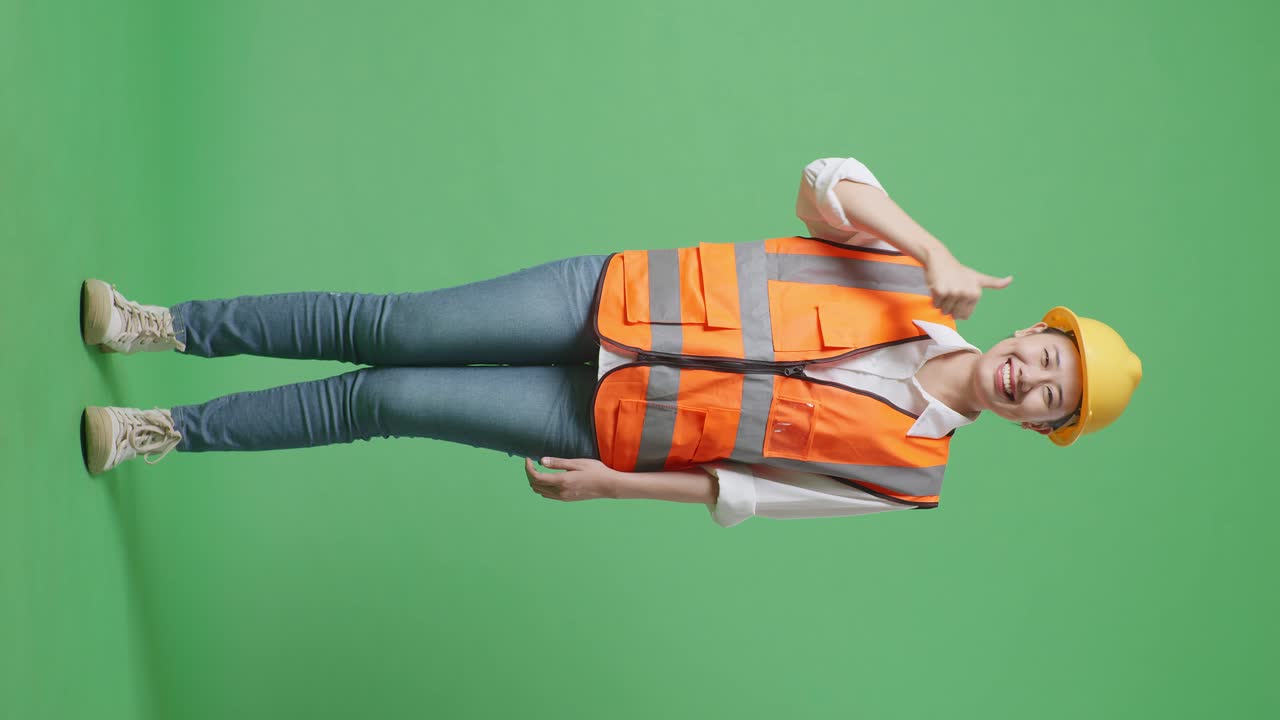 안전 헬을 입은 아시아 여성 엔지니어가 미소 짓고 엄지손가락을 보여주며 녹색 화면 배경 스튜디오에 서 있는 동안 카메라에 제스처를 합니다.