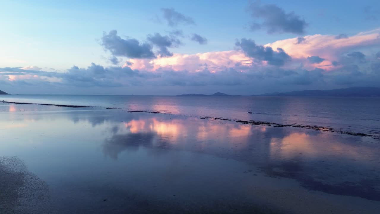 Drone footage of sunrise over Koh Phangan Thailand with a small boat moving across the calm tropical sea and the sunlight reflecting boat and clouds brightly on the water surface