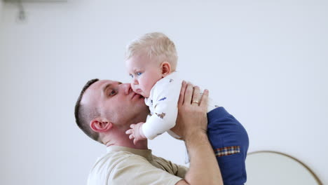 father is holding and kisses his beloved son. baby boy is in a bad mood and pushes away from dad. white backdrop.