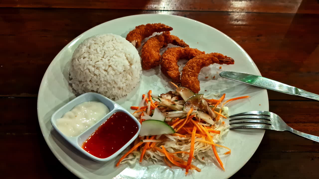 A push-in close-up shot of a delicious plate of crispy breaded fried shrimp, served with white rice, fresh salad, and dipping sauces at a restaurant in Gili Meno, Indonesia