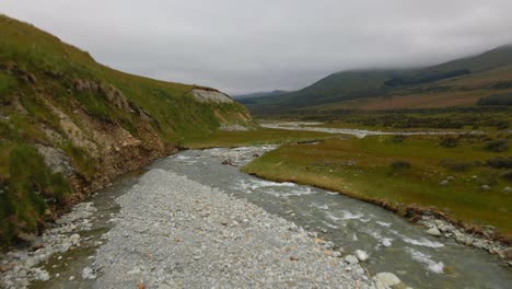 Rocky-riverbed-next-to-steep-grassy-slope-in-the-lush-hills-of-Canterbury,-New-Zealand-on-a-cloudy-day