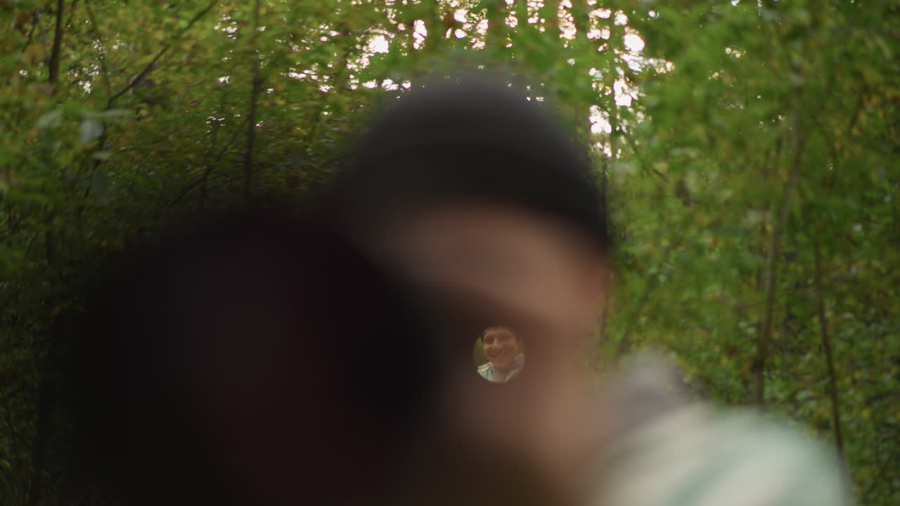 Die Nahaufnahme einer Gewehrlaufperspektive zeigt einen Mann beim Zielen. Die dunkle Mündung rahmt ein winziges, fokussiertes Fadenkreuz in der Mitte ein, eine verschwommene Mütze und Schultern im Vordergrund, dichte grüne Bäume und ein sanfter Bokeh-Hintergrund, ein Scout inspiziert das Zielfernrohr