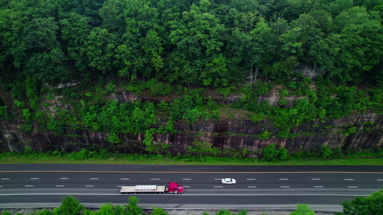 Aerial drone tracking shot of a red flatbed semi truck hauling cargo along I-24 through Monteagle’s forested cliffs, highlighting logistics.
