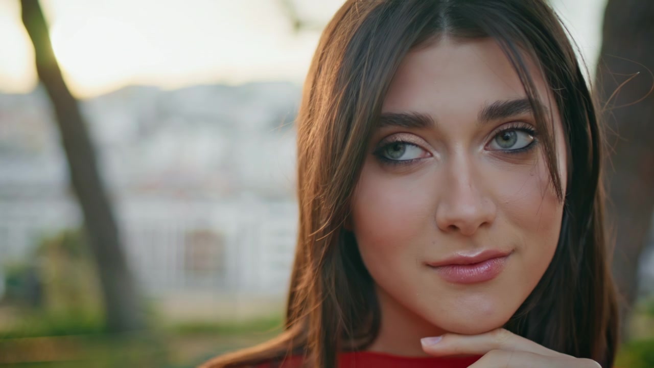 Hermosa mujer tocándose la cara en primer plano. Chica soñadora sonriendo en un parque de la ciudad al atardecer.