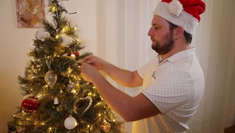 Man-wearing-festive-Santa-hat-decorating-traditional-Christmas-tree-arranging-lights-and-decorations-in-the-evening