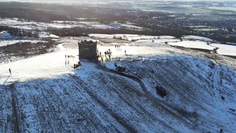 Snowy-Rivington-Pike-tower-Winter-hill-summit-aerial-view-people-sledding-downhill-at-sunrise-pull-back-reveal