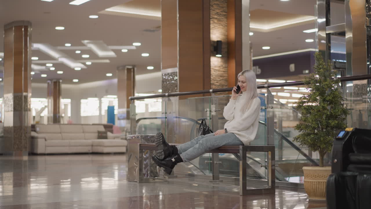 graceful lady seated on bench in shopping mall holding smartphone to ear laughing during call with legs crossed wearing chunky boots and denim jeans amidst glittering retail lights, blurred shoppers