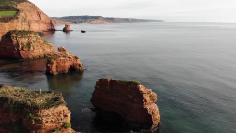 Cliffside-rocks-covered-with-grass-in-shallow-water-with-seagulls-flying-over,-camera-rises-to-reveal-the-coastline-at-Ladram-Bay