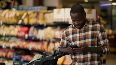 Ein-Mann-Mit-Schwarzer-Hautfarbe-Im-Karierten-Hemd-Tippt-Und-Chattet-Auf-Einem-Schwarzen-Telefon,-Neben-Ihm-Steht-Ein-Einkaufswagen-In-Einem-Supermarkt