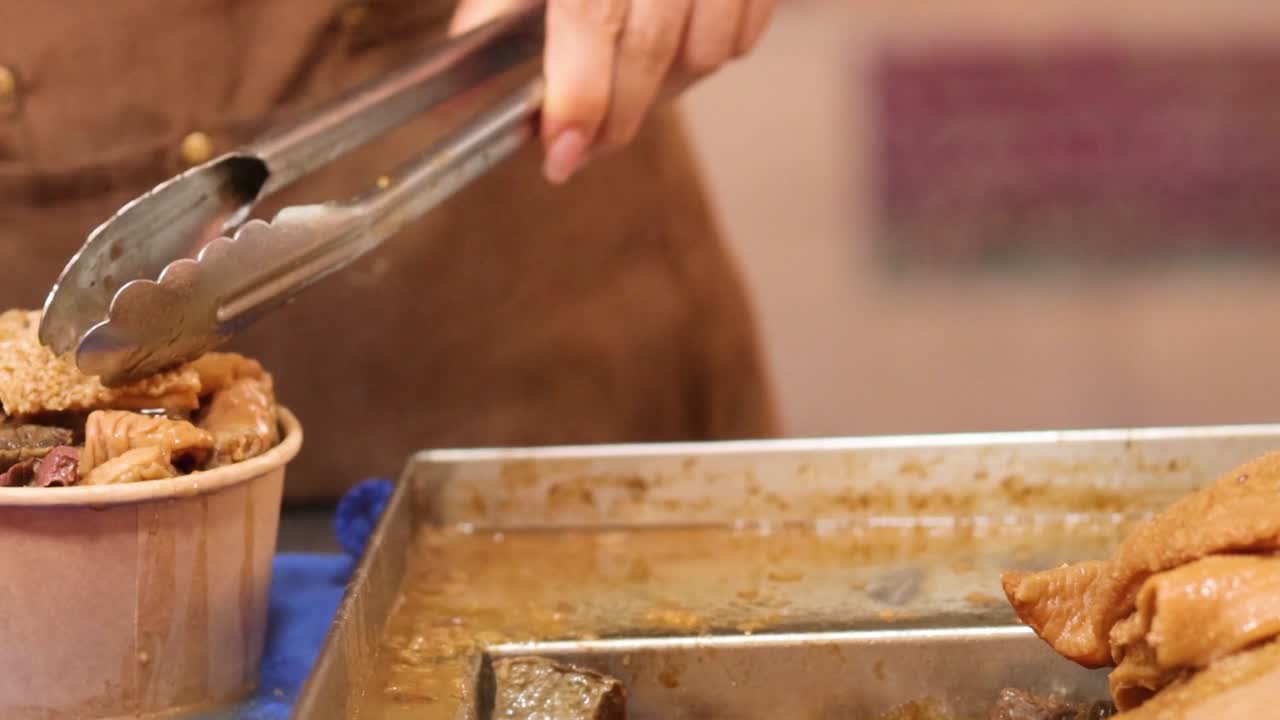 Close-up of hands using tongs and ladle to serve steaming meat into a paper bowl.
