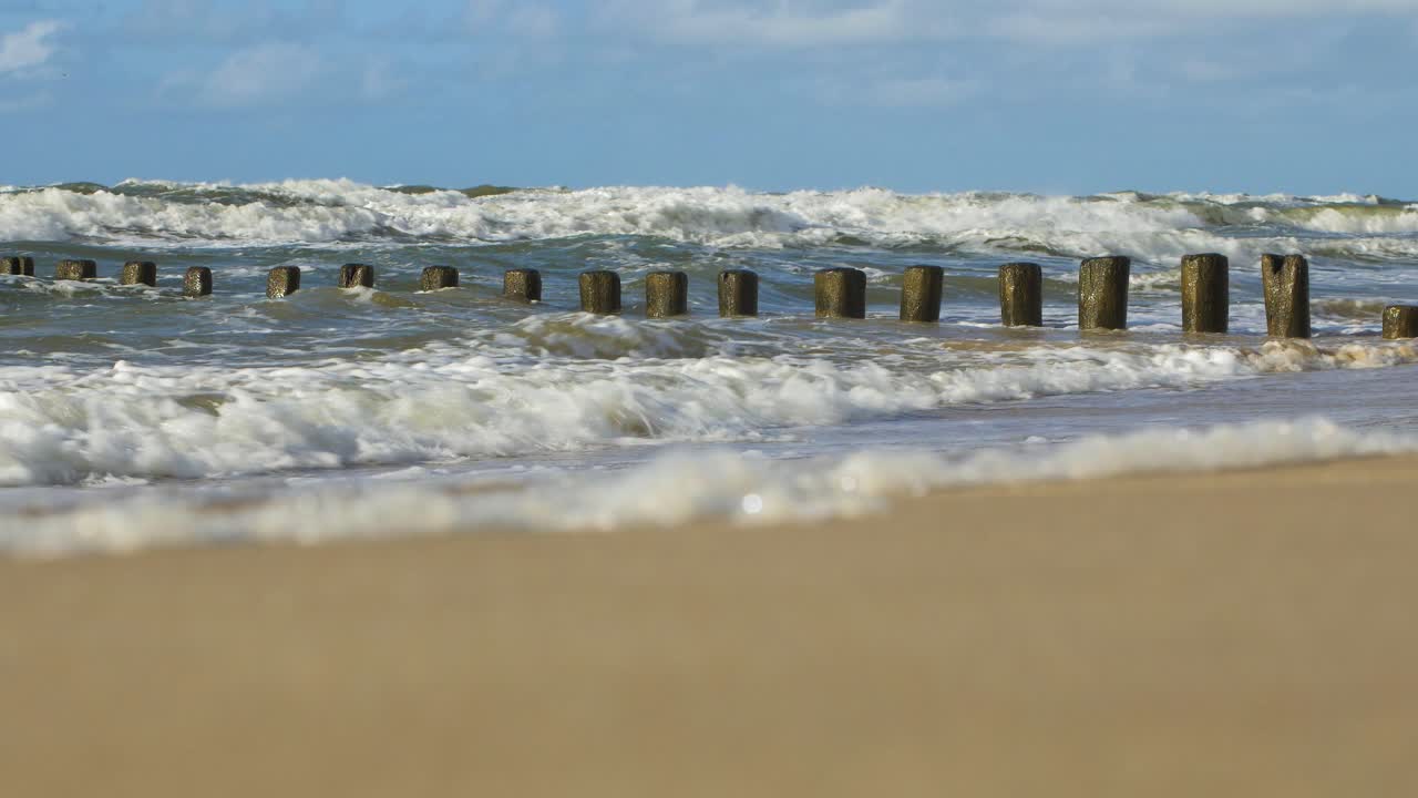 Stormy waves breaking against old wooden pier on the beach, white sand coast, sunny day, Baltic sea, distant low angle medium shot