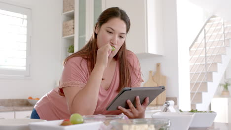 happy plus size biracial woman making meal in kitchen using tablet and eating fruit, slow motion