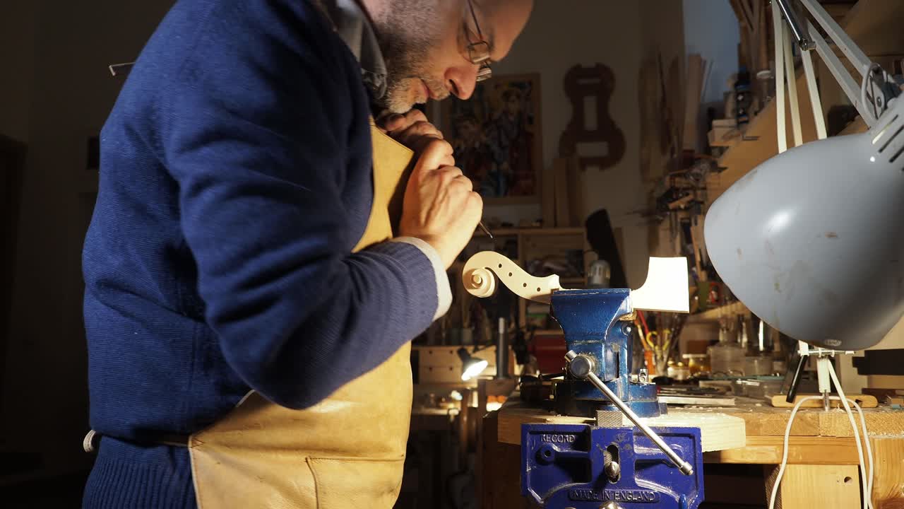 Master luthier leans over his workbench carving the scroll of a violin by hand under bright lamplight, the focused posture and warm tones capture the dedication artistry of traditional violin making