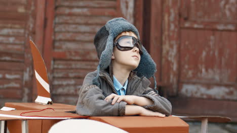 portrait of a cute little boy with red hair in hat and glasses sitting outdoor at wooden model of plane and smiling at the camera