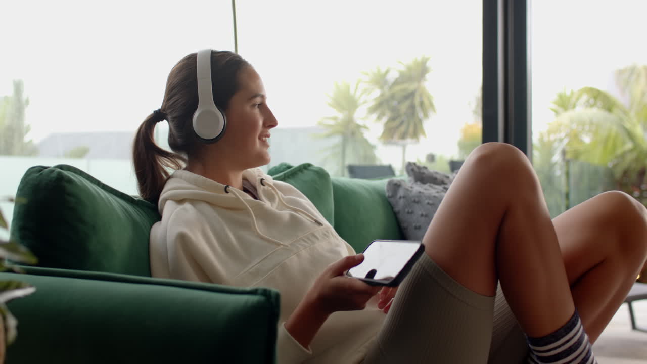 Relaxing on couch, woman wearing headphones and using smartphone, enjoying music, at home