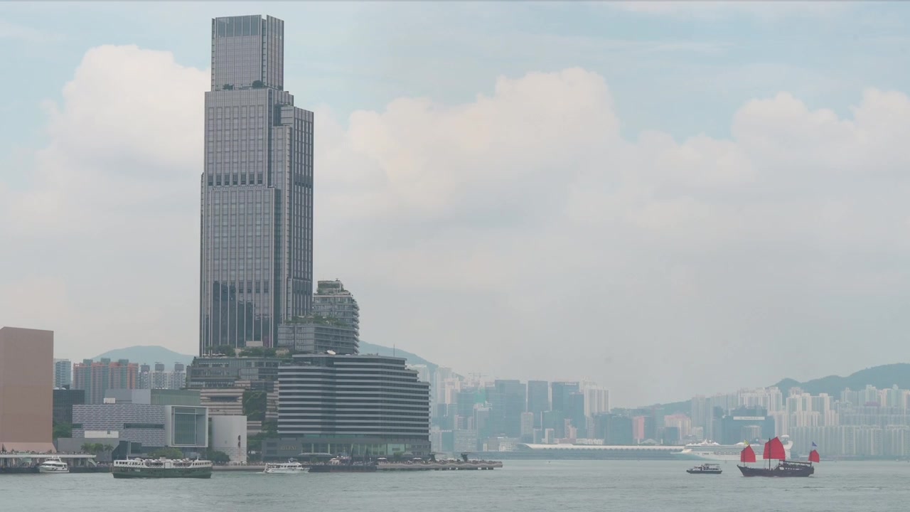 Um barco junco de velas vermelhas, modelado a partir de antigos navios chineses e agora uma atração turística, desliza pelo Porto Victoria ao lado de outras embarcações, com o moderno horizonte de Kowloon ao fundo.