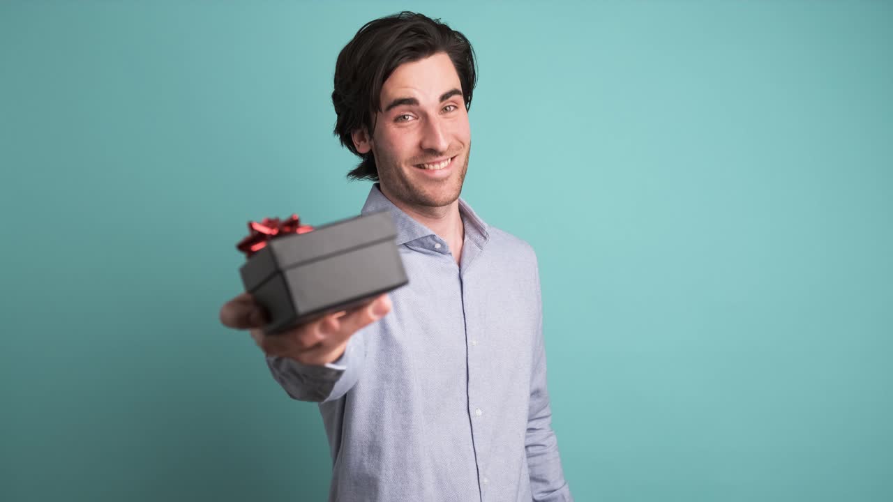 Happy man giving gift box and looking at camera in blue studio