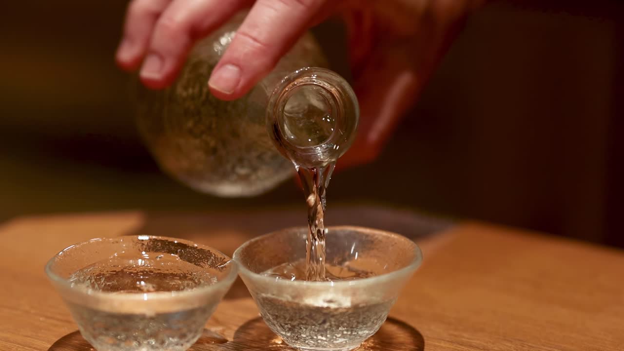 Close-up of a hand pouring liquid from a glass bottle into two small glass cups on a wooden surface.