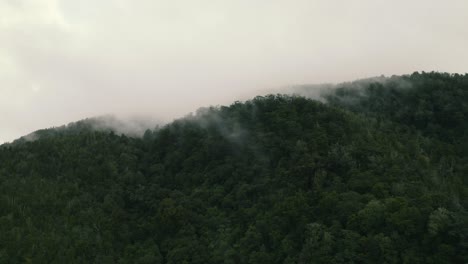 Long-aerial-zooming-out-shot-of-the-top-of-a-mountain-covered-by-clouds-and-fog-in-west-coast-New-Zealand