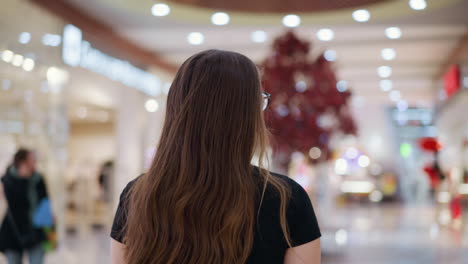 rear view of a woman with long flowing hair and glasses walking through a brightly lit mall, blurry figure in the background adds depth to the scene