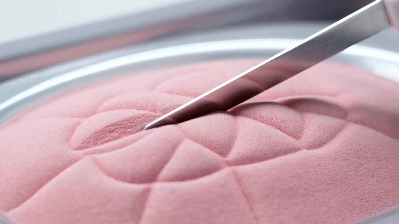 Una spatola in acciaio inossidabile preme e raschia su polvere rosa pastello testurizzata o schiuma vellutata all'interno di un piatto rotondo poco profondo, creando impronte allungate, strisce appiattite e riflessi tenui.