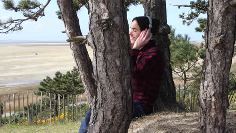 shot of an authentic hipster man listening to music with headphones whilst sitting in the sun against a tree trunk